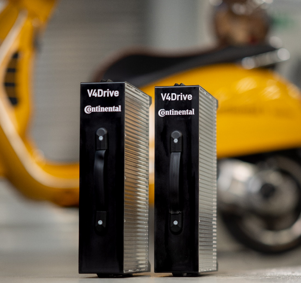 Two black V4Drive battery modules by Continental stand upright on a surface. A blurred yellow scooter is in the background, adding a contrasting color to the scene. The handles on the front of the battery modules are visible, emphasizing their portability.